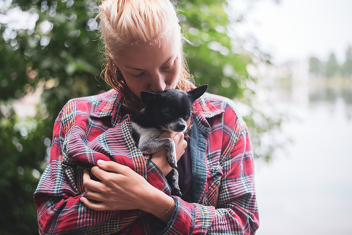 blonde woman by lake and green trees kisses small chihuahua inside top