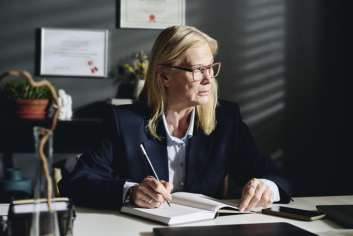 Thoughtful blond psychologist sitting at desk with diary in office