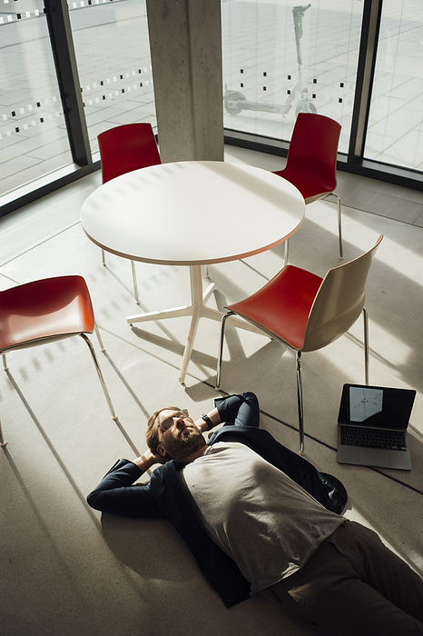 Businessman lying on floor with laptop and chairs in office