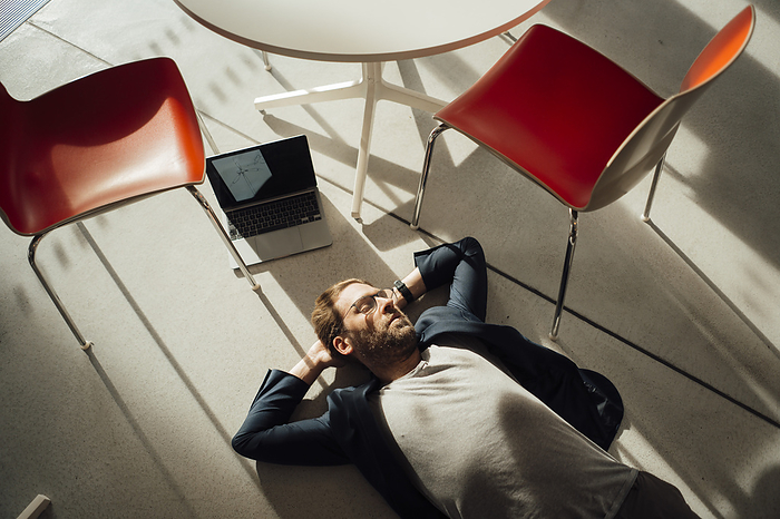 Businessman relaxing on floor with laptop and chairs in office