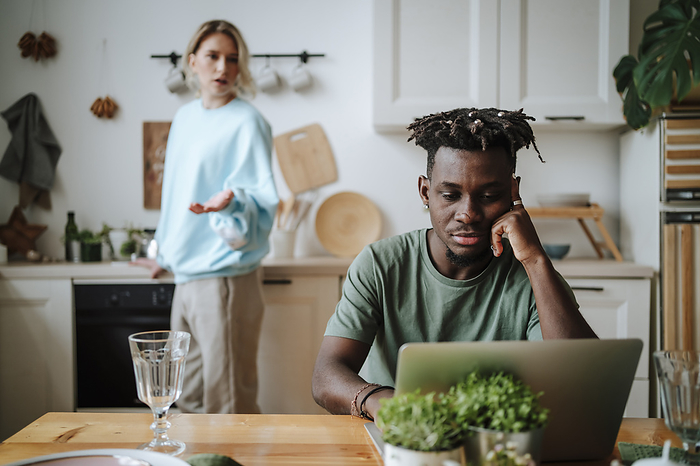 Stressed man watching laptop with woman arguing in background at home