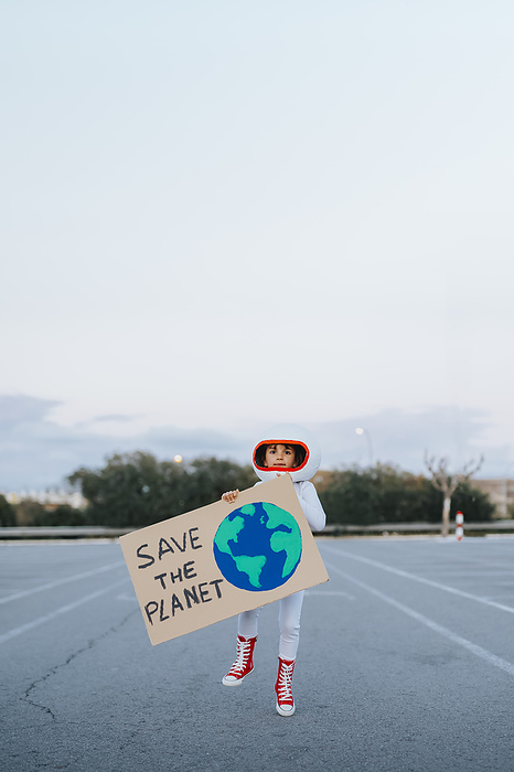 Astronaut girl holding Save The Planet placard standing in parking lot Astronaut girl holding Save The Planet placard standing in parking lot