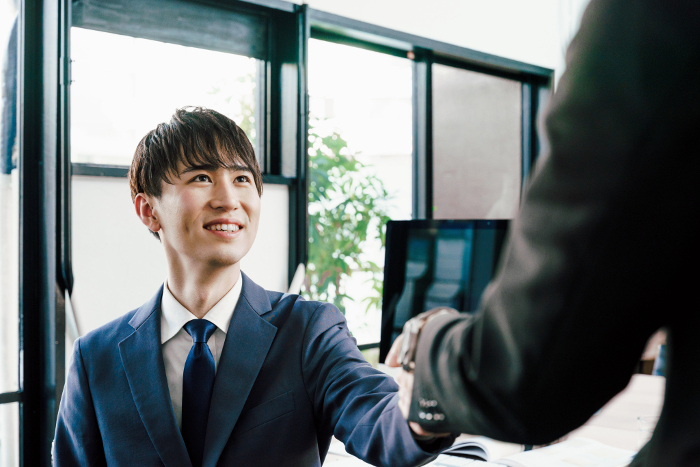 A young Japanese businessman in his 20s in a suit shaking hands in front of a computer monitor in his office (People)