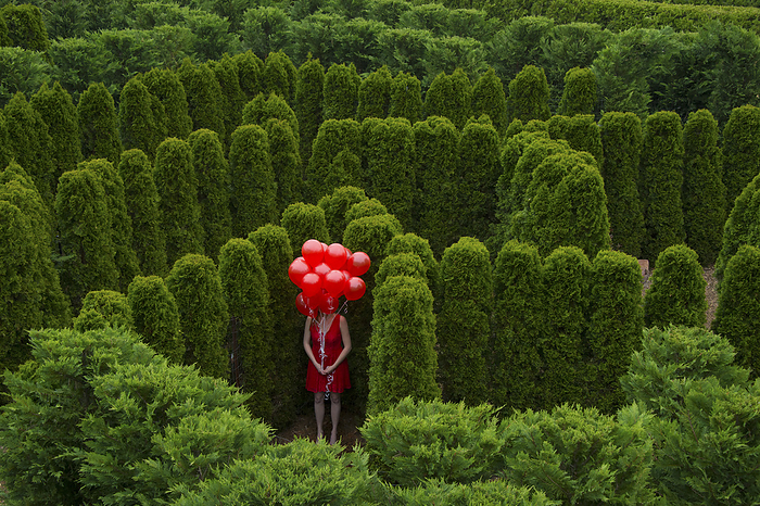 Young woman stands with a cluster of red balloons obscuring her face in a garden area; Luray, Virginia, United States of America, by Joel Sartore Photography / Design Pics