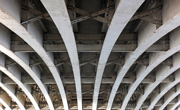 perspective view of curved arch shaped steel girders under an old road bridge with rivets and struts painted grey perspective view of curved arch shaped steel girders under an old road bridge with rivets and struts painted grey