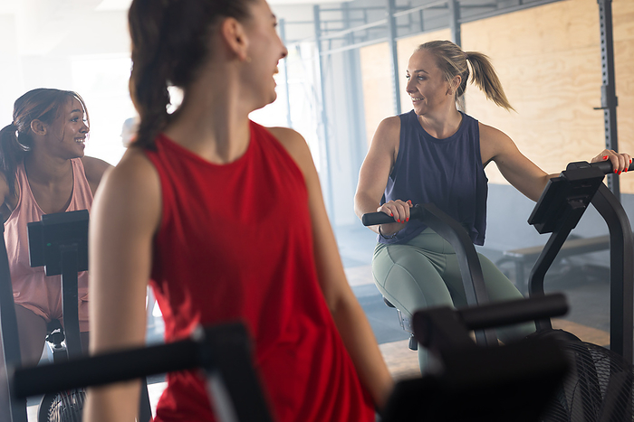 Tired multiracial women talking while exercising on elliptical trainer machine in health club. Unaltered, togetherness, cross training, gym, exercise, fitness and healthy lifestyle concept.