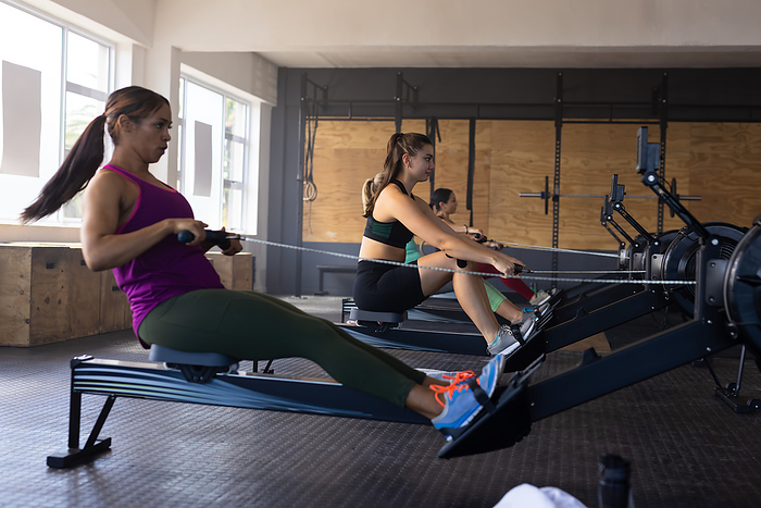 Side view of diverse dedicated female friends exercising on rowing machines in health club. Copy space, unaltered, togetherness, cross training, exercise, gym, fitness and healthy lifestyle.