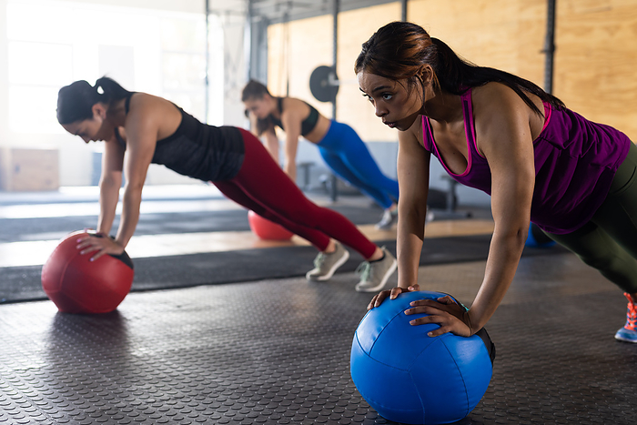 Multiracial women with fitness balls practicing push-ups in health club, copy space. Unaltered, togetherness, cross training, exercise, fitness, gym, effort, sunlight and healthy lifestyle concept.