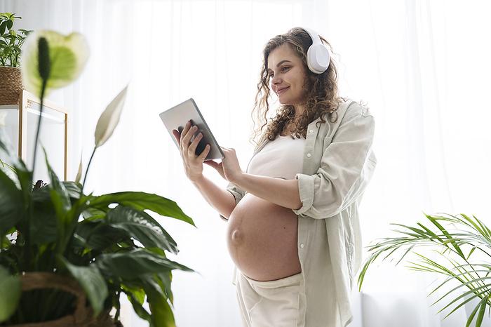 Smiling pregnant woman with headphones using tablet PC at home