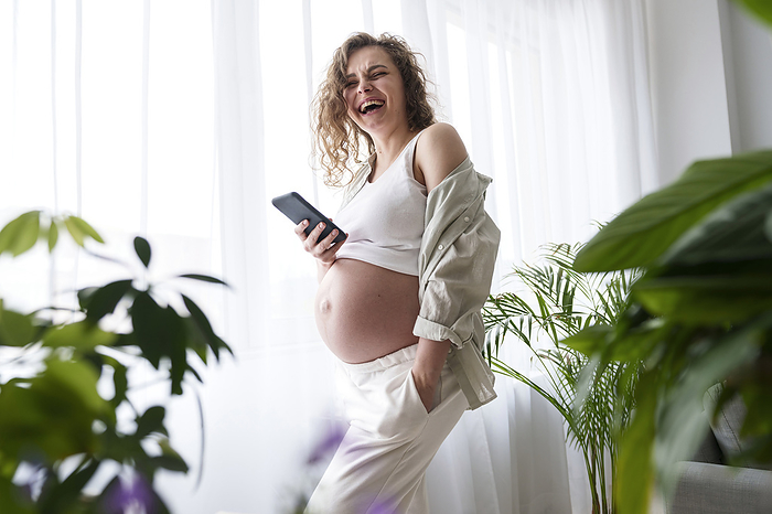 Happy pregnant woman with hand in pocket holding smart phone at home