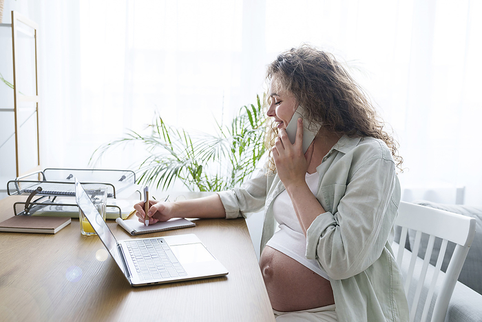 Pregnant freelancer writing and talking on smart phone at home office