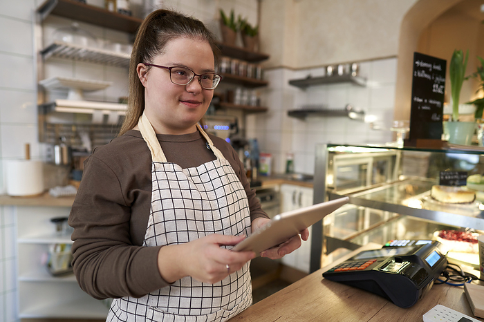 Caucasian woman with down syndrome browsing digital tablet while working in a cafe Smiling cafe owner with down syndrome standing with tablet PC