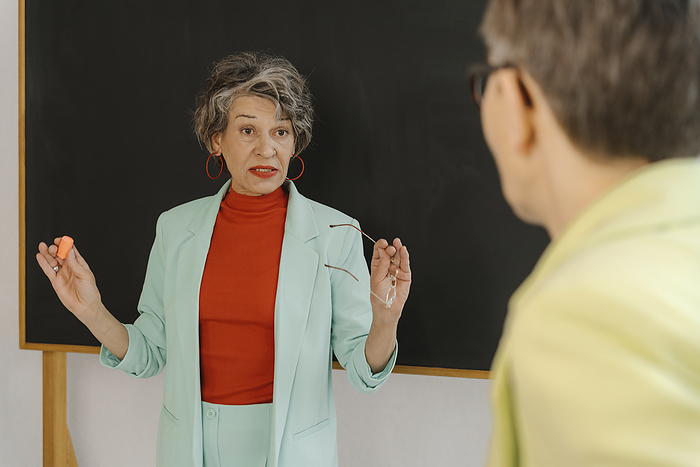 Woman in a bright suit standing in front of blackboard explaining something to another woman.