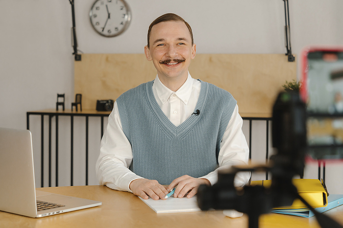 Smiling man sitting in classroom looking at camera recording online class