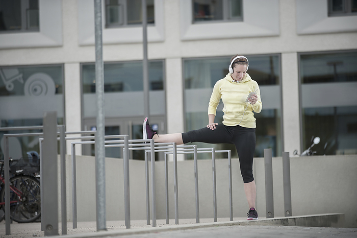young woman streches herself in urban enviroment, functional fitness,urban sports, fitness, freetime,sportive lifestyle Young woman stretching her leg on railings and listening to music, Bavaria, Germany