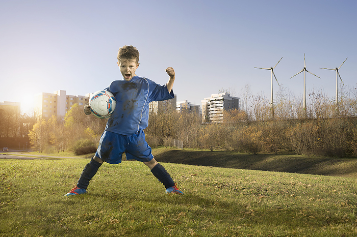 young football player cheers in clean urban enviroment with wind wheels,save enviorment, no polution, Dirty soccer player cheering on field and wind turbines with city in background, Bavaria, Germany