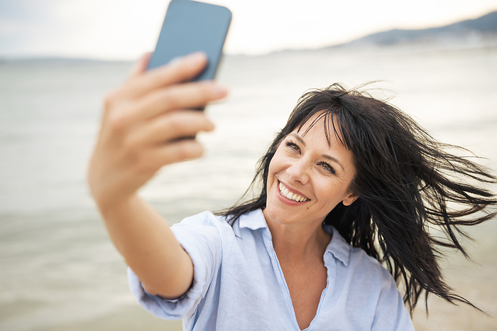 Happy woman with tousled hair taking selfie through smart phone at beach