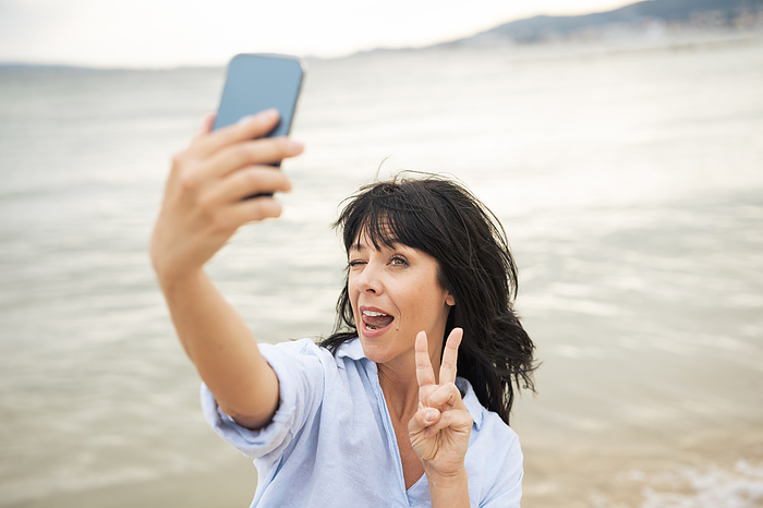 Woman making peace sign taking selfie through smart phone at beach