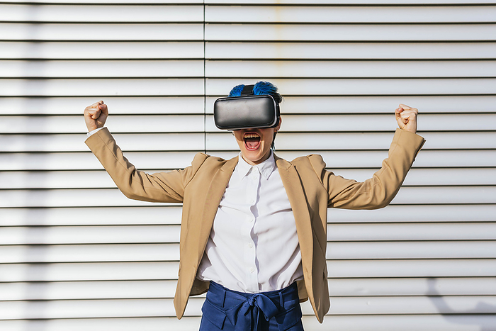 Cheerful businesswoman wearing virtual reality headset flexing muscles in front of wall
