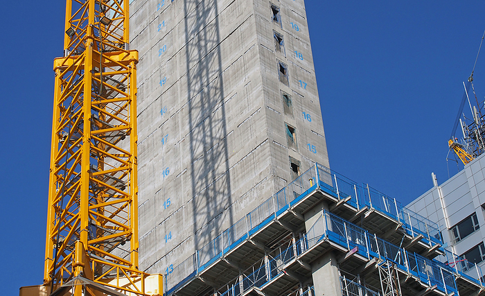close up of a large urban construction site with a yellow tower crane casting a shadow on a large concrete building and safety fences against a blue sky close up of a large urban construction site with a yellow tower crane casting a shadow on a large concrete building and safety fences against a blue sky