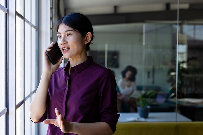 Happy asian casual businesswoman standing next to window in office talking on smartphone, copy space. Work, business, communication and casual office, unaltered.