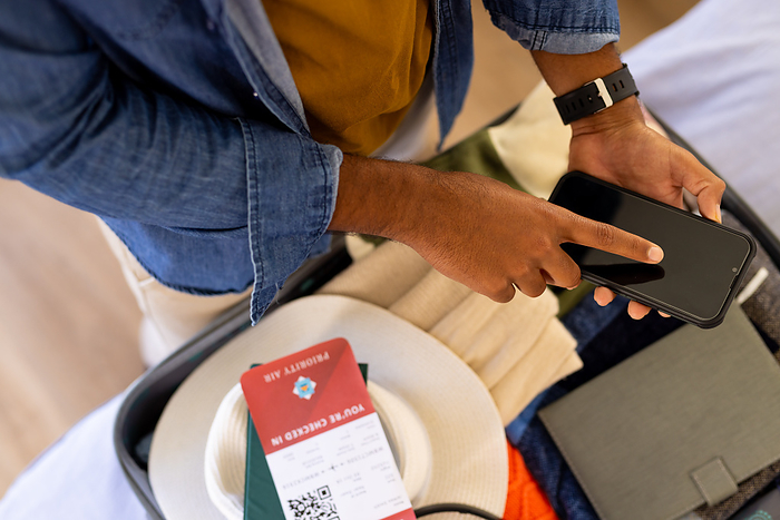 Midsection of biracial man with packed suitcase checking flight boarding pass using smartphone. Travel, vacations, preparation, communication, free time and lifestyle.