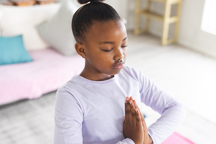 African american girl doing yoga and meditating in living room. Relaxation, childhood, active lifestyle and domestic life, unaltered.