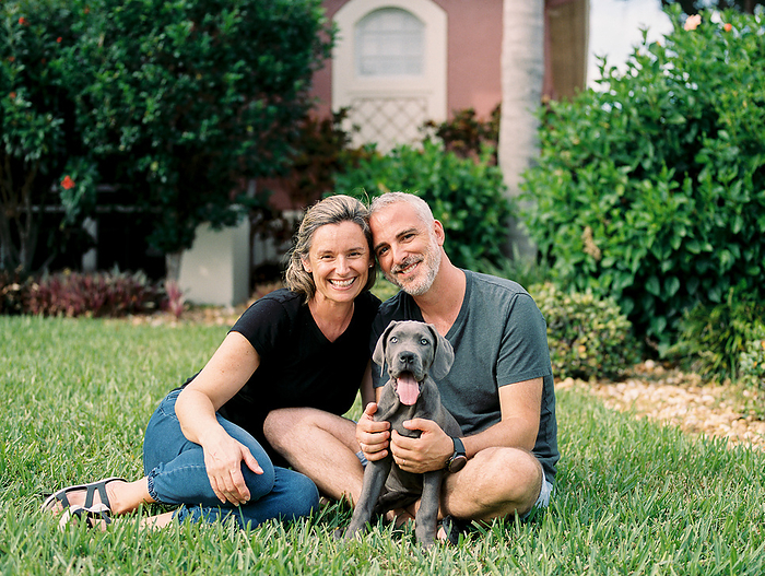 hispanic couple sits in their front yard, smiling with their puppy