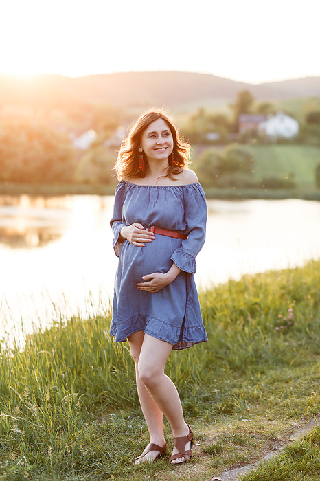 pregnant woman in blue dress holds her belly under rays of sun
