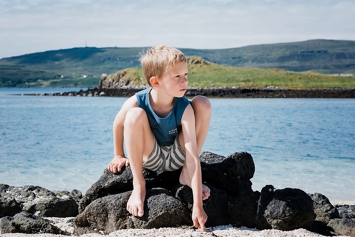 boy sat on a rock at the beach playing with the sand