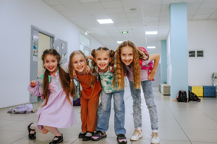 Joyful schoolgirls with arm around standing in school corridor