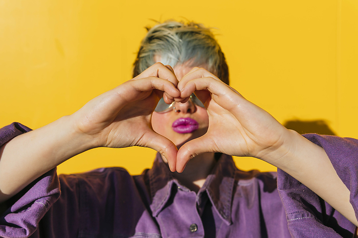 Woman making heart gesture and puckering in front of yellow wall