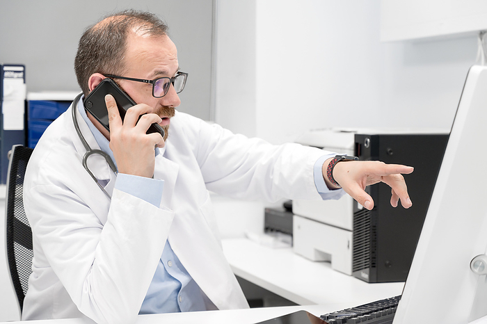 Male doctor pointing with finger on desktop computer while talking on the phone, discussing treatment with colleague. Male doctor pointing with finger on desktop computer while talking on the phone, discussing treatment with colleague., by Zoonar DAVID HERRAEZ