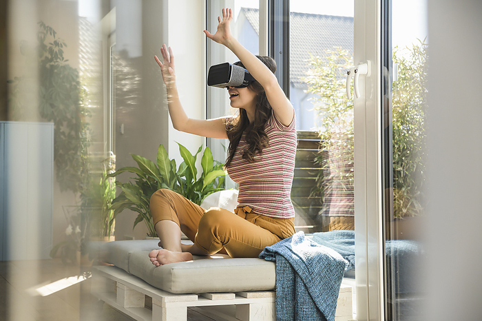 Young woman sitting at the window at home wearing VR glasses