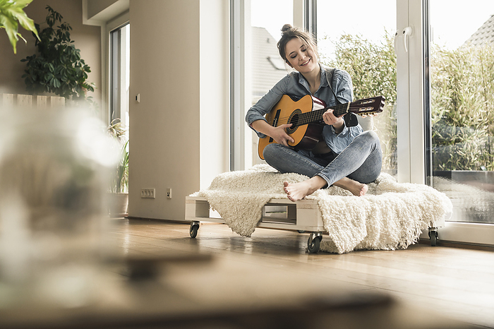 Smiling woman sitting at the window at home playing guitar