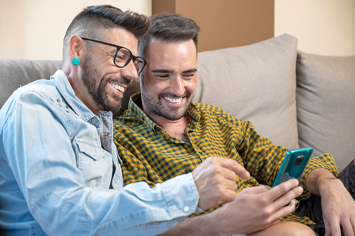Happy young gay couple using mobile phone while sitting on a sofa in the living room Happy young gay couple using mobile phone while sitting on a sofa in the living room, by Zoonar DAVID HERRAEZ
