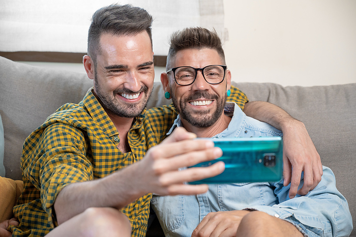 Cheerful gay couple sitting on the couch hugging and looking at the mobile phone, watching a movie, scrolling through the social network Cheerful gay couple sitting on the couch hugging and looking at the mobile phone, watching a movie, scrolling through the social network, by Zoonar DAVID HERRAEZ