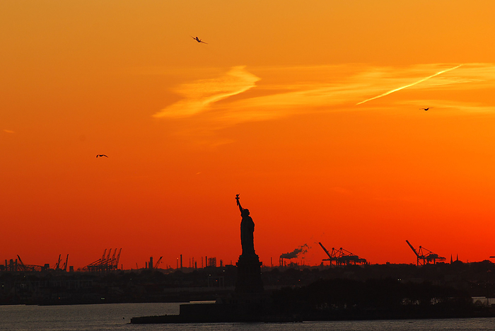 Silhouette of Statue of Liberty and Liberty Island in New York against an orange sunset sky Silhouette of Statue of Liberty and Liberty Island in New York against an orange sunset sky, by Zoonar Raffael Herrm