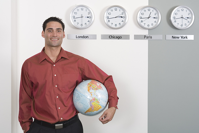 Businessman Holding Globe Standing by Clocks Showing International Time Zones, by Albert C. Karges / Design Pics