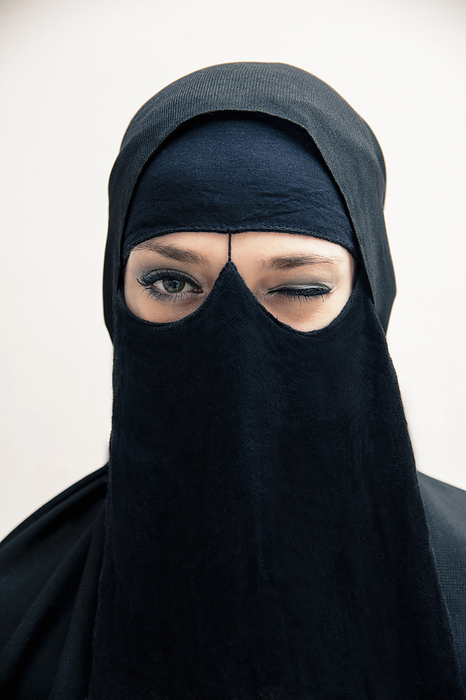 Close-up portrait of young woman wearing black, muslim hijab and muslim dress, winking and looking at camera, eyes showing eye makeup, studio shot on white background, by Bettina Salomon / Design Pics