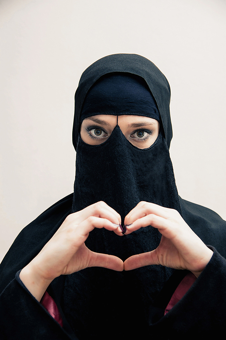 Close-up portrait of young woman wearing black, muslim hijab and muslim dress, making heart shape with hands, looking at camera, eyes showing eye makeup, studio shot on white background, by Bettina Salomon / Design Pics