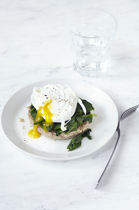 Poached egg with soft yolk and spinach on an english muffin, on plate with fork and glass of water, studio shot on white background, by Jodi Pudge / Design Pics