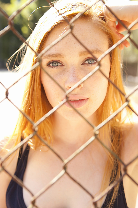Close-up portrait of young woman standing behind chain link fence in park near the tennis court on a warm summer day in Portland, Oregon, USA, by Ty Milford / Design Pics