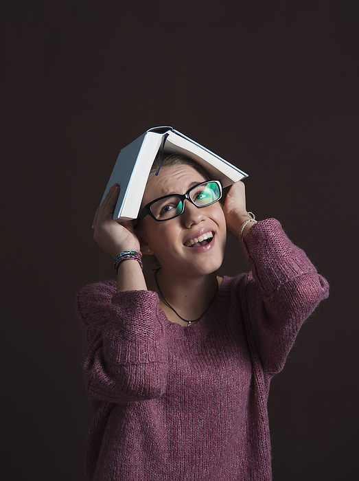 Portrait of Teenage Girl wearing Eyeglasses, holding Open Book over Head with Anxious Expression, Studio Shot on Black Background, by Uwe Umstätter / Design Pics