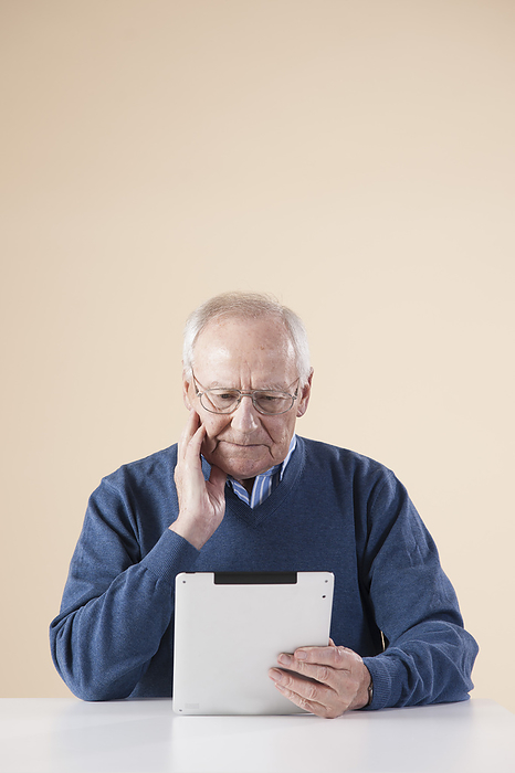 Senior Man Sitting at Table, Looking at Tablet PC, Studio Shot on Beige Background, by Uwe Umstätter / Design Pics