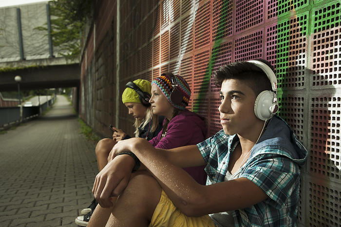 Children sitting next to wall outdoors, wearing headphones and listening to music, Germany, by Uwe Umstätter / Design Pics