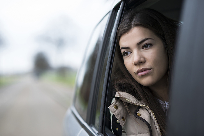 Portrait of young woman sitting inside car and looking out of window and day dreaming on overcast day, Germany, by Uwe Umstätter / Design Pics