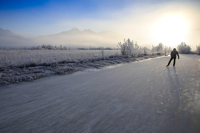 View taken from behind of a Caucasian man backcountry ice skating, nordic blading, into the winter sun on a partly sunny, partly foggy, winter day with frost covered grass and trees lining Rabbit Slough, with Pioneer Peak, Twin Peaks, and the Chugach Mountains in the background in Wasilla; Alaska, United States of America, by Amber Johnson / Design Pics
