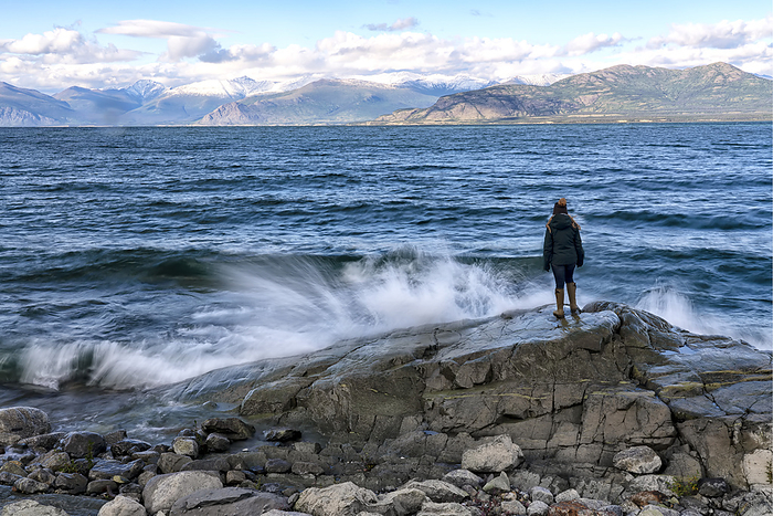 View taken from behind of a woman standing on a rock while the waves crah into the shore of Kluane Lake on a windy day; Yukon, Canada, by Robert Postma / Design Pics