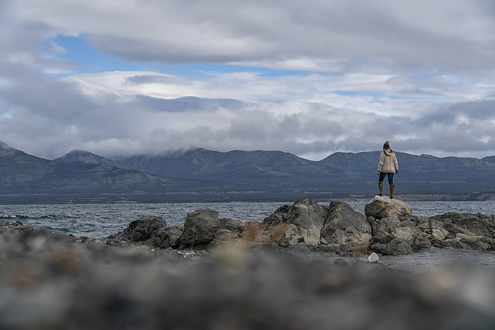 View taken from behind of a woman standing along the rocky shore of Kluane Lake; Yukon, Canada, by Robert Postma / Design Pics